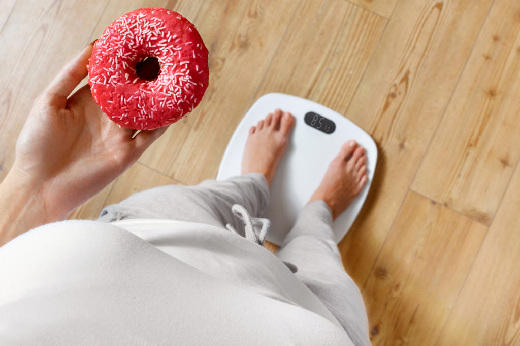Concept of overweight - a woman holding a doughnut while standing on a scale
